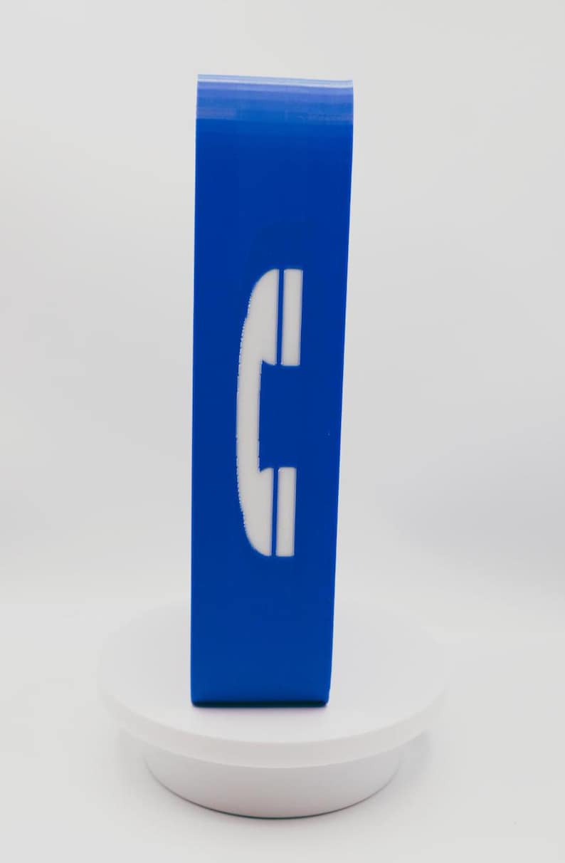 May include: A rectangular blue block featuring a white telephone handset icon centered on the front face. The object stands upright on a circular white base, displaying a smooth, solid-colored surface with a clear, high-contrast graphic symbol.