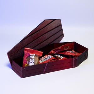 Puede incluir: Una caja en forma de ata&uacute;d burdeos con tapa abierta, llena de caramelos envueltos individualmente. La caja tiene un acabado texturizado y es de madera. Los caramelos est&aacute;n en envoltorios rojos y marrones, con la palabra "UNREAL" visible.