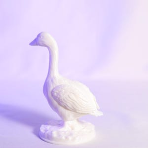 May include: A white ceramic goose figurine with a long neck and detailed feather textures. The goose stands on a textured base, set against a soft, gradient background. The figurine is a decorative item.