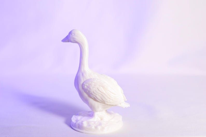 May include: A white ceramic goose figurine with a long neck and detailed feather textures. The goose stands on a textured base, set against a soft, gradient background. The figurine is a decorative item.