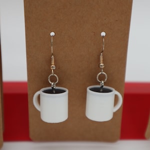May include: A pair of white coffee mug earrings with black coffee inside. The earrings are hanging from gold hooks.
