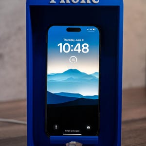 May include: A bright blue, rectangular phone stand shaped like a classic telephone booth with the word Phone embossed in white at the top. A smartphone is docked inside, displaying a digital clock, date, and a mountain landscape wallpaper.