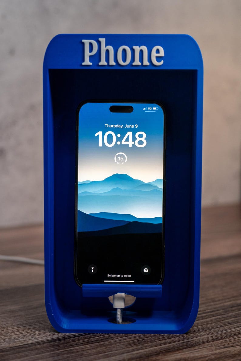 May include: A bright blue, rectangular phone stand shaped like a classic telephone booth with the word Phone embossed in white at the top. A smartphone is docked inside, displaying a digital clock, date, and a mountain landscape wallpaper.