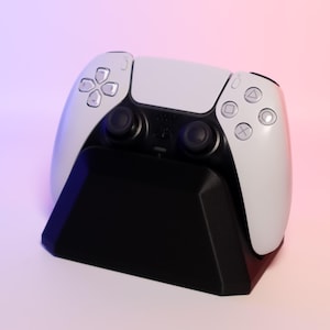 May include: A white video game controller with black buttons and a black charging stand. The controller is on the stand and the stand is on a white surface.