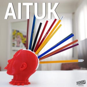 May include: A red, 3D-printed head-shaped pencil holder with various colored pencils inserted. The word "AITUK" is displayed in white block letters above the head. The pencils are yellow, red, blue, and brown. The base of the holder has a spiked collar.