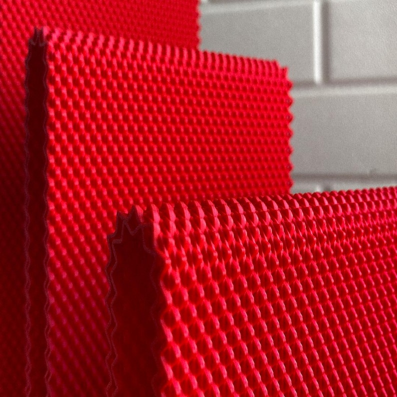 May include: Close-up of several layers of bright red, textured fabric. The fabric has a repeating pattern of small, raised dots, creating a grid-like appearance. The edges of the fabric are visible, showing a slightly wavy cut. The background is a blurred white.