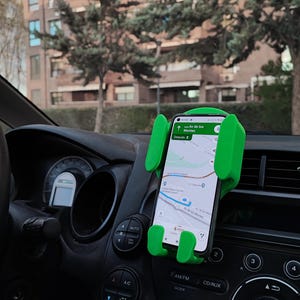 May include: A green phone holder for a car dashboard. The holder has two arms that extend to hold a phone in place. The phone is showing a map app with directions to Av de los Montes. The phone holder is attached to the dashboard of a car.