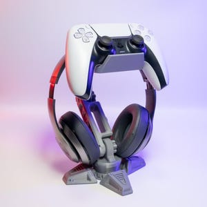 Op de afbeelding: Een grijze en witte gaming-headsetstandaard met een witte PlayStation 5-controller die aan de bovenkant is bevestigd. De standaard heeft een stervormige basis.