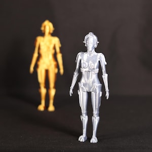 Może przedstawiać: Srebrna figurka robota o szczegółowym wzornictwie. Robot ma ludzkie ciało z ruchomymi kończynami i głowę z twarzą. Robot stoi na czarnym tle.