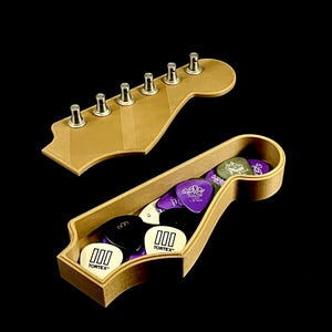 Puede incluir: Una caja de madera con forma de clavijero de guitarra con tapa, llena de p&uacute;as de guitarra. Las p&uacute;as son negras, blancas y moradas, con el texto "000 TORTEX" en algunas. La tapa tiene seis clavijas de afinaci&oacute;n plateadas.