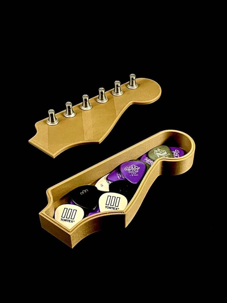 Puede incluir: Una caja de madera con forma de clavijero de guitarra con tapa, llena de p&uacute;as de guitarra. Las p&uacute;as son negras, blancas y moradas, con el texto "000 TORTEX" en algunas. La tapa tiene seis clavijas de afinaci&oacute;n plateadas.