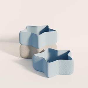 Puede incluir: Tres recipientes en forma de estrella de estilo minimalista. Dos son de color azul claro y uno beige. Los recipientes tienen una superficie texturizada y están apilados. Los recipientes están vacíos.