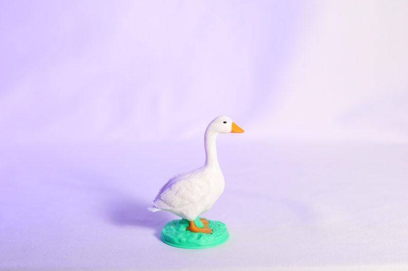 May include: A white goose figurine with an orange beak and feet stands on a small, round, green base. The goose has a long neck and is set against a soft purple background.