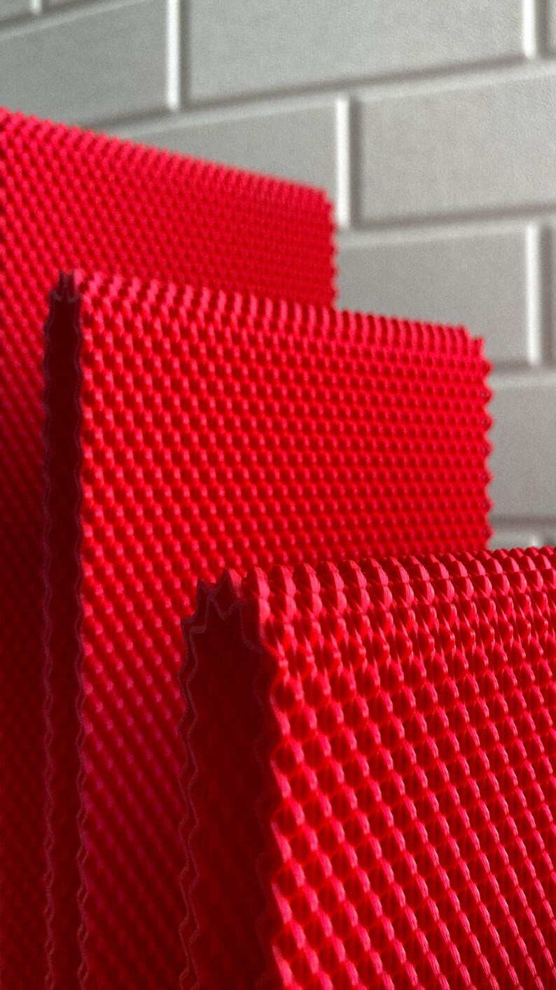 May include: Close-up of three red textured rectangular mats with a repeating pattern of small, raised bumps. The mats are stacked, revealing a textured edge. The background is a light grey, brick-like wall.