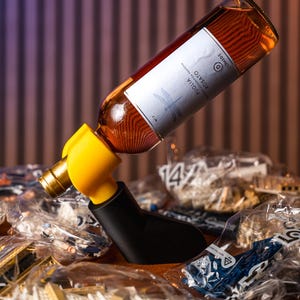 May include: A wine bottle is held by a black and yellow bottle holder. The bottle is filled with a light amber liquid and has a white label. The holder is resting on a surface covered with plastic bags.