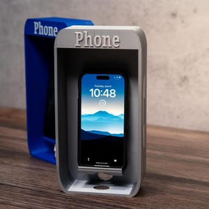 May include: A gray phone stand shaped like a vintage telephone booth with the word Phone embossed on the top. A smartphone with a blue mountain landscape wallpaper is resting inside the stand. A second blue phone booth stand is visible behind it.