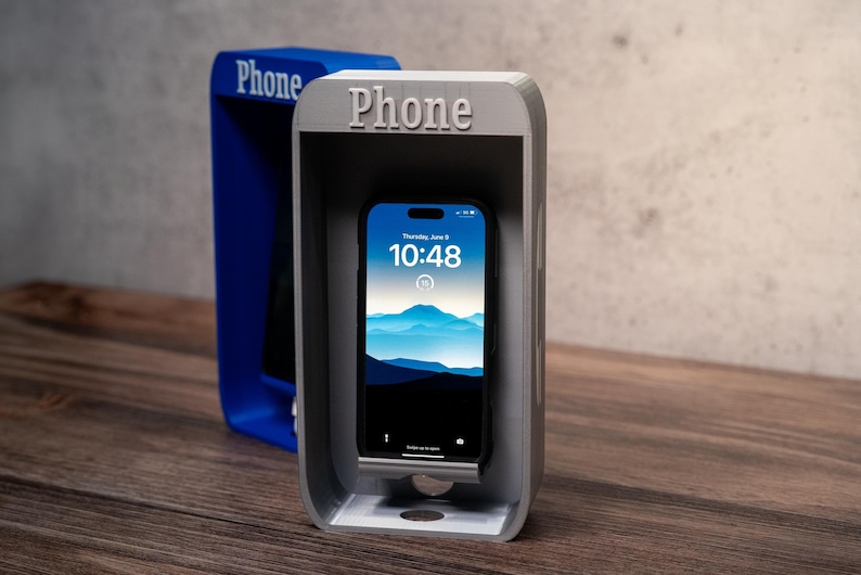 May include: A gray phone stand shaped like a vintage telephone booth with the word Phone embossed on the top. A smartphone with a blue mountain landscape wallpaper is resting inside the stand. A second blue phone booth stand is visible behind it.