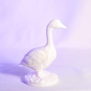 May include: A white ceramic goose figurine. The goose is standing upright with its head held high. The figurine has detailed feather textures and stands on a textured base. The background is a gradient of purple and white.
