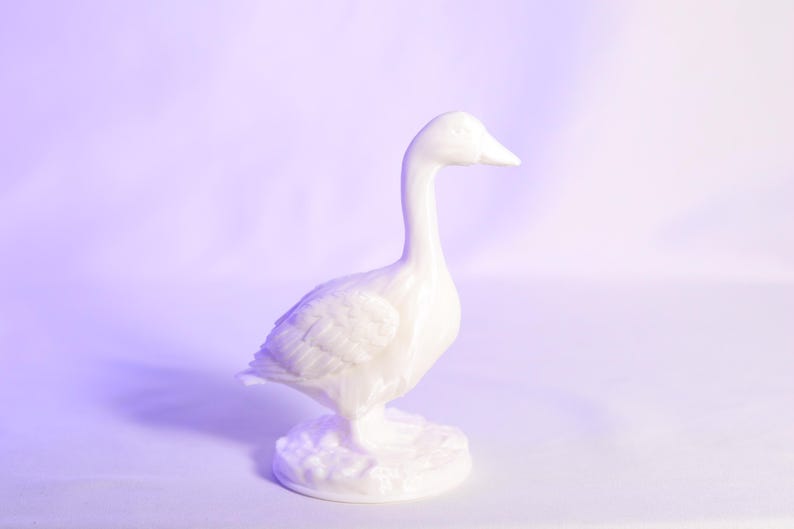 May include: A white ceramic goose figurine. The goose is standing upright with its head held high. The figurine has detailed feather textures and stands on a textured base. The background is a gradient of purple and white.