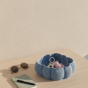 Puede incluir: Un cuenco azul acanalado en forma de nube contiene llaves y un llavero rosa. Gafas de sol redondas y un pequeño cuaderno verde con un bolígrafo descansan sobre una superficie de madera clara. El fondo es una pared beige neutra.