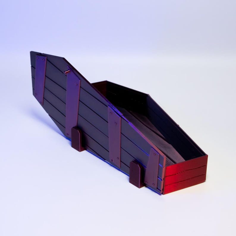 Puede incluir: Una caja de madera oscura con una tapa inclinada y detalles en rojo. La caja tiene forma rectangular con listones de madera verticales y est&aacute; sostenida por dos peque&ntilde;os pies rectangulares. La caja est&aacute; vac&iacute;a y se encuentra sobre una superficie blanca.