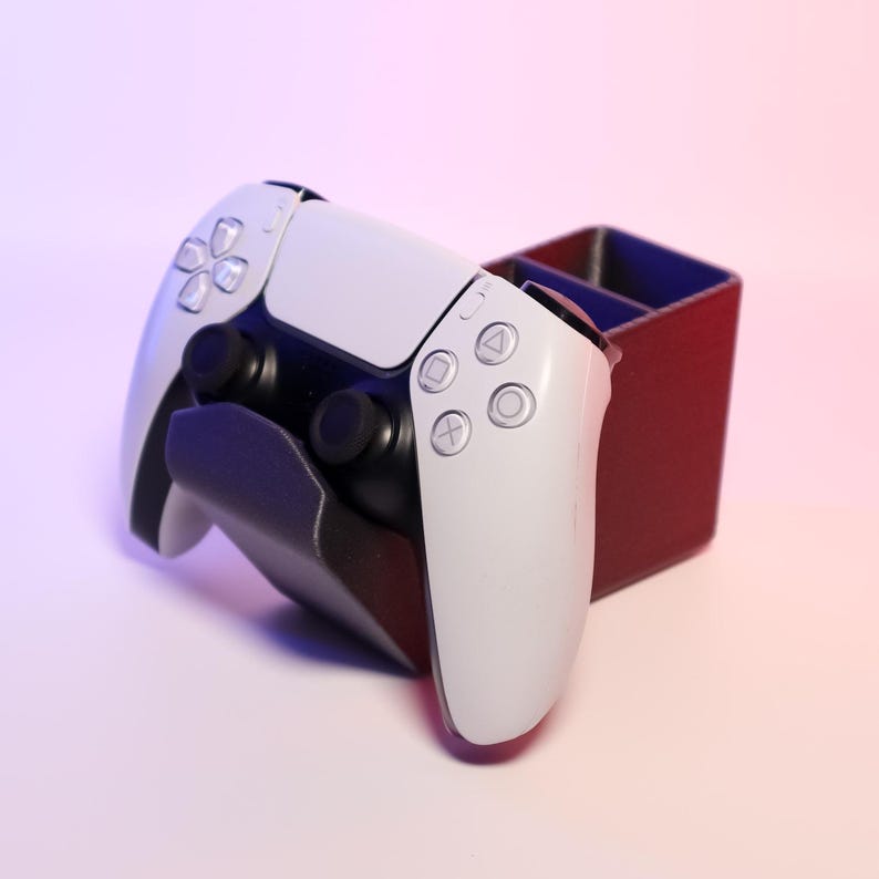 May include: A white PlayStation 5 controller is resting in a black and red charging stand. The controller has black buttons and a white faceplate.