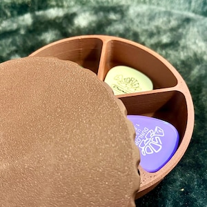 Puede incluir: Un soporte circular marrón para púas de guitarra con tres compartimentos, parcialmente abierto. En el interior hay púas de guitarra verdes y moradas. El soporte está sobre una superficie verde texturizada.