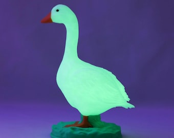 Könnte beinhalten: Eine leuchtend grüne Gänsefigur mit orangefarbenem Schnabel und Füßen. Die Gans steht auf einem grünen, grasartigen Sockel. Der Hintergrund ist ein Farbverlauf von Lila und Blau.