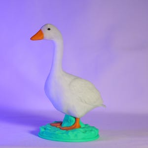 以下が含まれることがあります： オレンジ色のくちばしと足を持つ白いガチョウの置物。緑色の台座の上に立っています。ガチョウは長い首をしており、紫色の背景に配置されています。装飾品です。