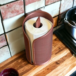 May include: A brown paper towel holder with a woven texture, holding a roll of white paper towels. The holder has a curved design and is mounted on a wall. The kitchen accessory is on a wooden surface.