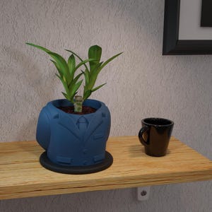 Puede incluir: Un macetero de cerámica azul con forma de chaqueta de traje con una planta verde creciendo en su interior. El macetero está colocado en una estantería de madera con una taza de café negra.