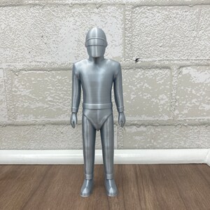 May include: A silver-colored figurine of a robot with a helmet and a futuristic design. The robot is standing on a wooden surface.
