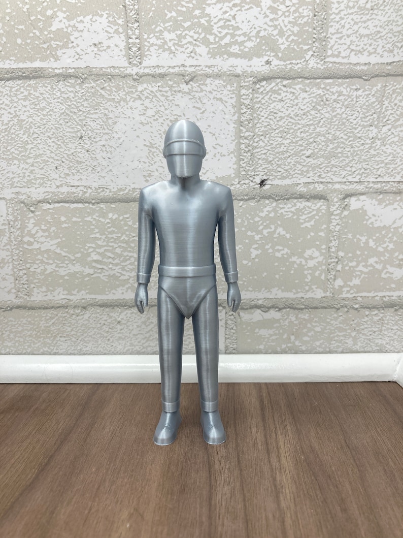 May include: A silver-colored figurine of a robot with a helmet and a futuristic design. The robot is standing on a wooden surface.