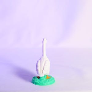 May include: A white goose figurine with a long neck, standing on a green base with orange accents. The figurine is set against a soft purple background, creating a whimsical and decorative piece.