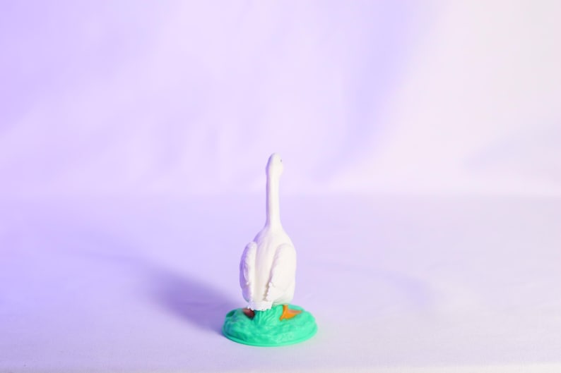 May include: A white goose figurine with a long neck, standing on a green base with orange accents. The figurine is set against a soft purple background, creating a whimsical and decorative piece.