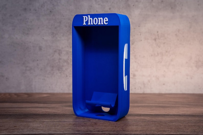 May include: A bright blue rectangular phone stand with rounded corners. The top features the white text "Phone" and the right side displays a white telephone handset icon. The interior includes a small cradle for holding a mobile device.