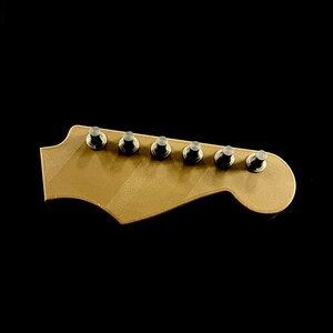 Puede incluir: Una pala de guitarra dorada con seis clavijas de afinaci&oacute;n plateadas. La pala tiene una forma cl&aacute;sica con un extremo curvado y una serie de l&iacute;neas diagonales. El fondo es negro liso.