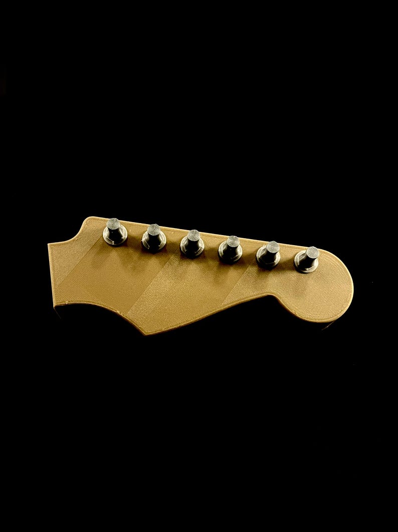 Puede incluir: Una pala de guitarra dorada con seis clavijas de afinaci&oacute;n plateadas. La pala tiene una forma cl&aacute;sica con un extremo curvado y una serie de l&iacute;neas diagonales. El fondo es negro liso.