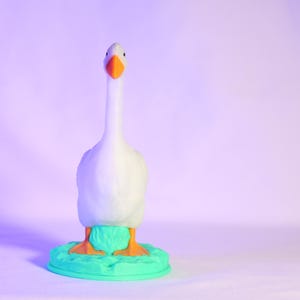 以下が含まれることがあります： オレンジ色のくちばしと足を持つ白いガチョウの置物。ターコイズ色の台座の上に立っています。ガチョウは長い首をしていて、正面を向いています。背景は紫と白のグラデーションです。