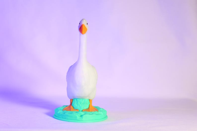 以下が含まれることがあります： オレンジ色のくちばしと足を持つ白いガチョウの置物。ターコイズ色の台座の上に立っています。ガチョウは長い首をしていて、正面を向いています。背景は紫と白のグラデーションです。
