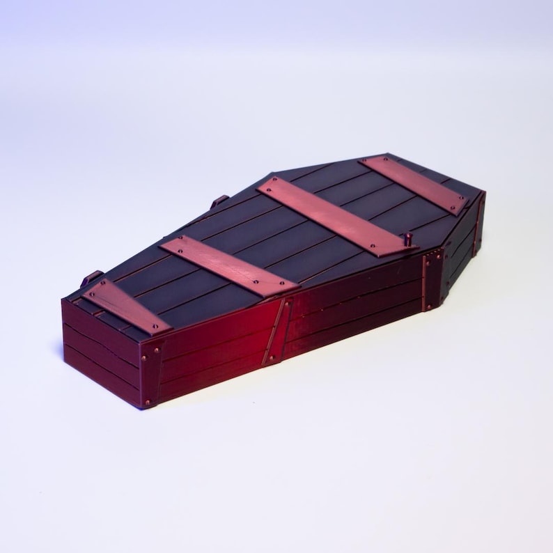 Puede incluir: Un ata&uacute;d en miniatura de madera oscura con detalles rojos. La tapa est&aacute; asegurada con tres correas de metal rojo. Los lados y la base tambi&eacute;n son rojos, creando un contraste llamativo. Un art&iacute;culo decorativo.