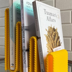 May include: A vibrant yellow book holder with a textured, wavy design, supporting three books. The books feature white covers and titles such as "Advanced 3D Printing" and "Txumari Alfaro". The holder is on a glass surface.