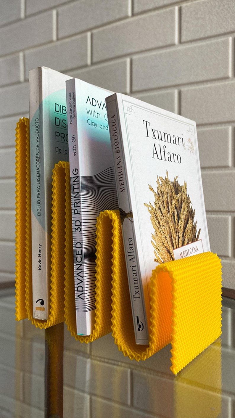 May include: A vibrant yellow book holder with a textured, wavy design, supporting three books. The books feature white covers and titles such as "Advanced 3D Printing" and "Txumari Alfaro". The holder is on a glass surface.