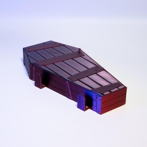 Puede incluir: Un peque&ntilde;o ata&uacute;d de madera oscura con detalles met&aacute;licos. El ata&uacute;d tiene forma rectangular con tapa inclinada. Est&aacute; construido con tablones de madera y bandas de metal, de color marr&oacute;n oscuro con toques rojos y morados.
