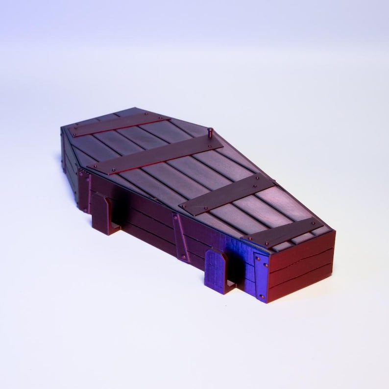 Puede incluir: Un peque&ntilde;o ata&uacute;d de madera oscura con detalles met&aacute;licos. El ata&uacute;d tiene forma rectangular con tapa inclinada. Est&aacute; construido con tablones de madera y bandas de metal, de color marr&oacute;n oscuro con toques rojos y morados.