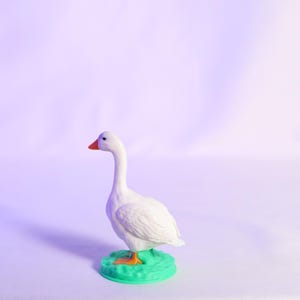 May include: A white goose figurine with an orange beak and feet stands on a green base. The goose is set against a soft, purple-toned background, creating a whimsical and charming aesthetic. This decorative item is perfect for home decor.