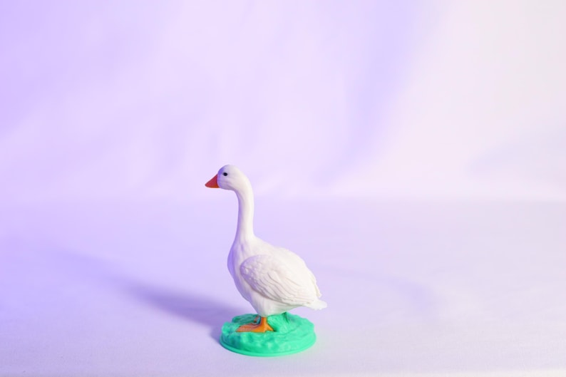 May include: A white goose figurine with an orange beak and feet stands on a green base. The goose is set against a soft, purple-toned background, creating a whimsical and charming aesthetic. This decorative item is perfect for home decor.