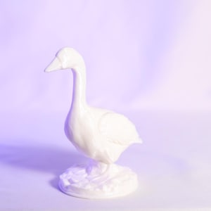 May include: A white ceramic goose figurine with a long neck and a detailed base. The goose is standing upright, with its head held high. The figurine is set against a soft purple background.