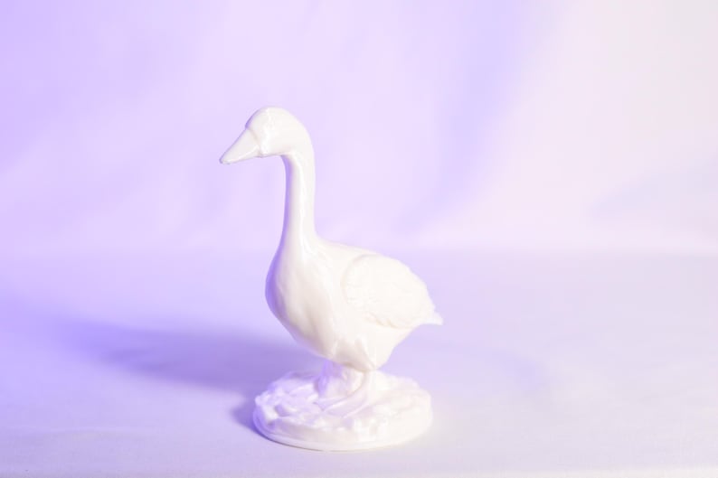 May include: A white ceramic goose figurine with a long neck and a detailed base. The goose is standing upright, with its head held high. The figurine is set against a soft purple background.