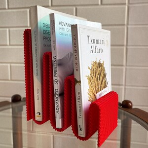 May include: A red, textured book holder with three books. The books have white covers and titles in Spanish and English. The holder is made of a flexible material and is designed to hold books upright. The books are on a glass surface.